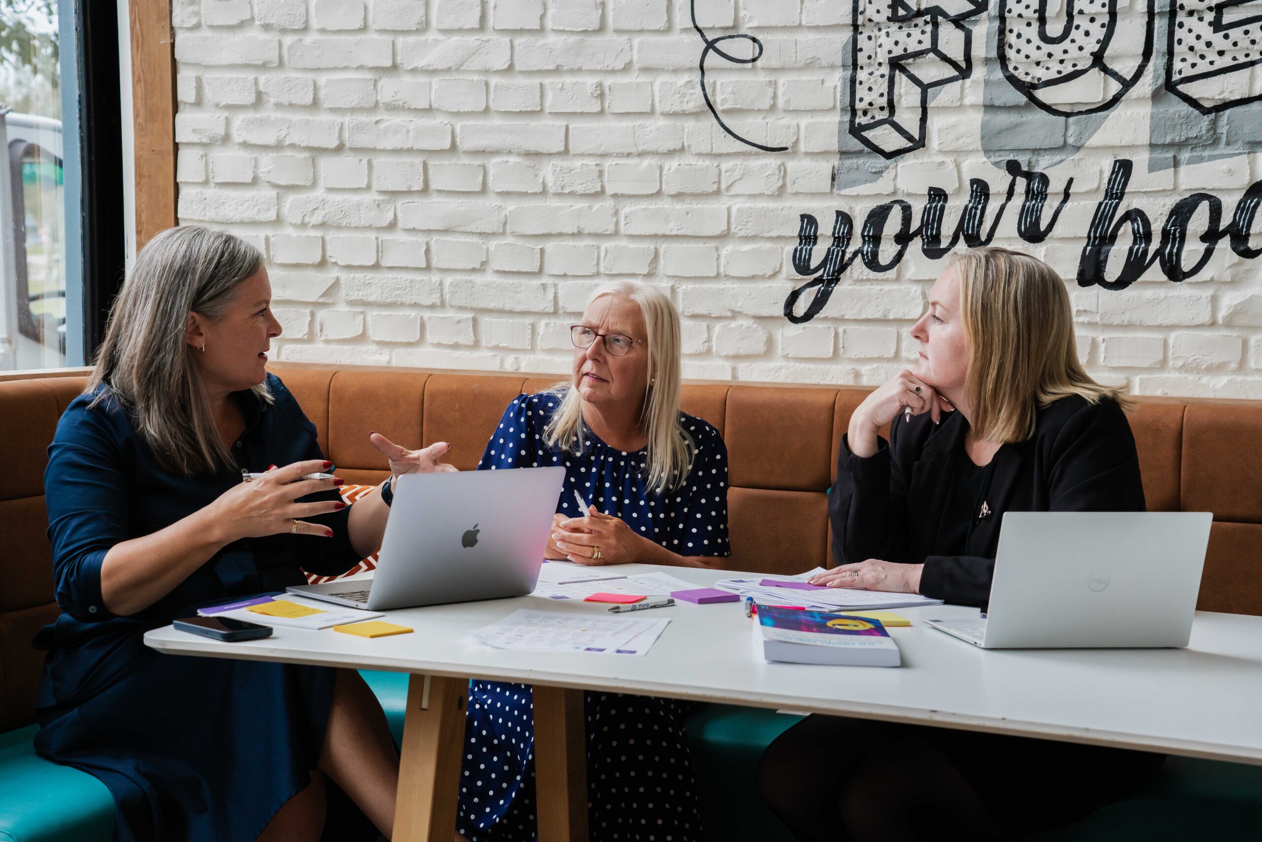 Three people, who happen to all be female, working with laptop, paper, post its and pens littering a modern office as three look interested in the discussion being had. 